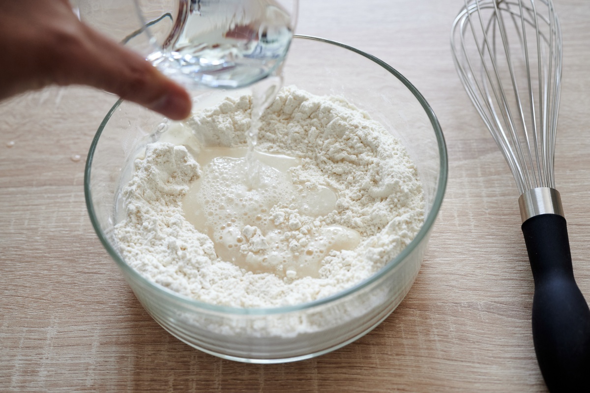 Eine Person gibt Wasser in eine Schüssel Mehl. Daneben liegrt ein Schneebesen zum Umrühren.
