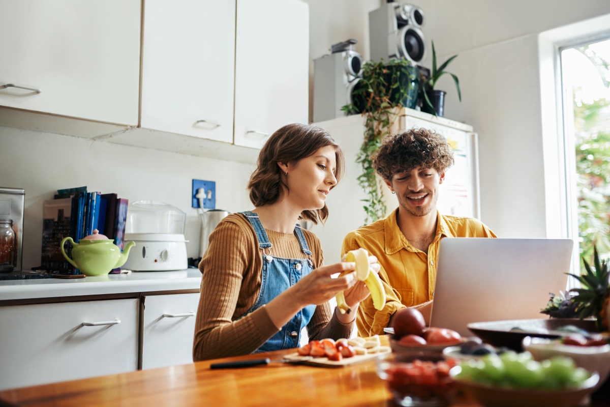 Paar bereitet gemeinsam Frühstück in der Küche zu während sie auf dem Laptop ihre Rezepte für die Woche planen.