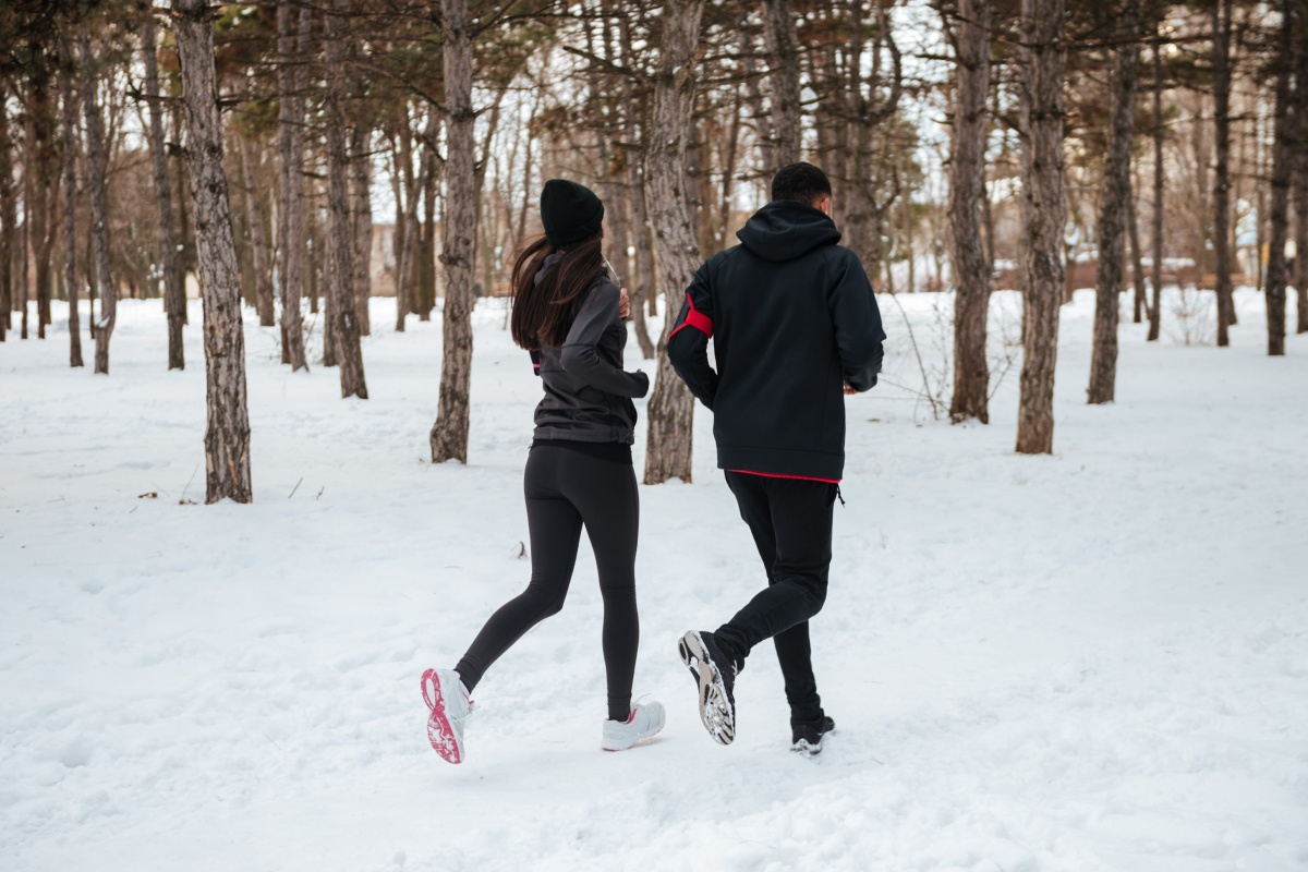 Ein Mann und eine Frau in dunkler Sportkleidung joggen durch einen verschneiten Wald.