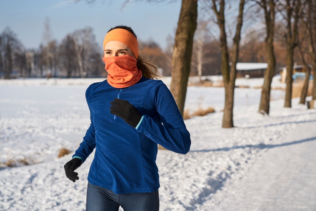 Frau joggt an einem sonnigen Wintertag durch verschneite Landschaft. Sie trägt Handschuhe, Stirnband und ein Halstuch, das sie bis über die Nase gezogen hat.