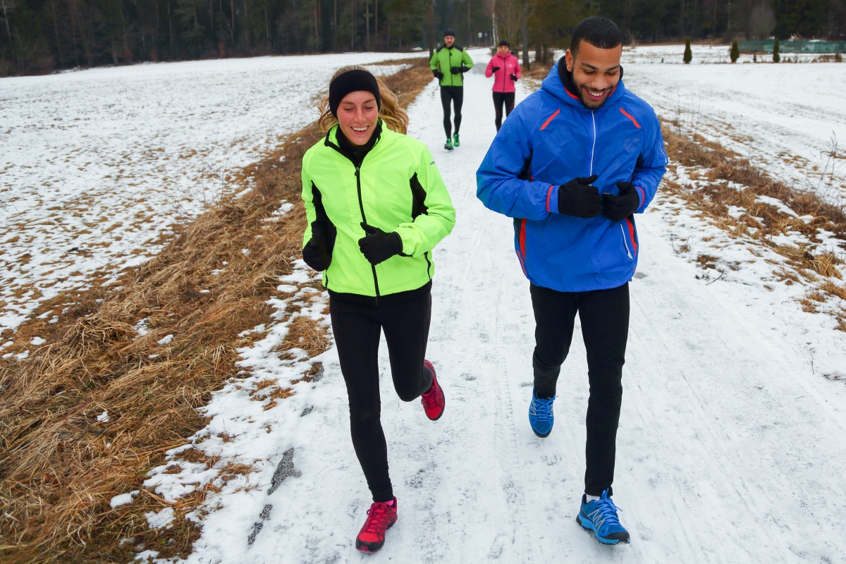 Laufgruppe joggt in bunter Sportkleidung auf verschneitem Weg durch ländliche Winterlandschaft.