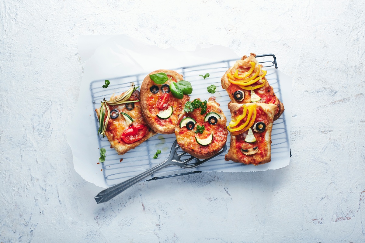 Mini-Pizzen in Gesicht-Form, dekoriert mit Gemüse und Oliven, auf einem Gitterrost mit Backpapier serviert.