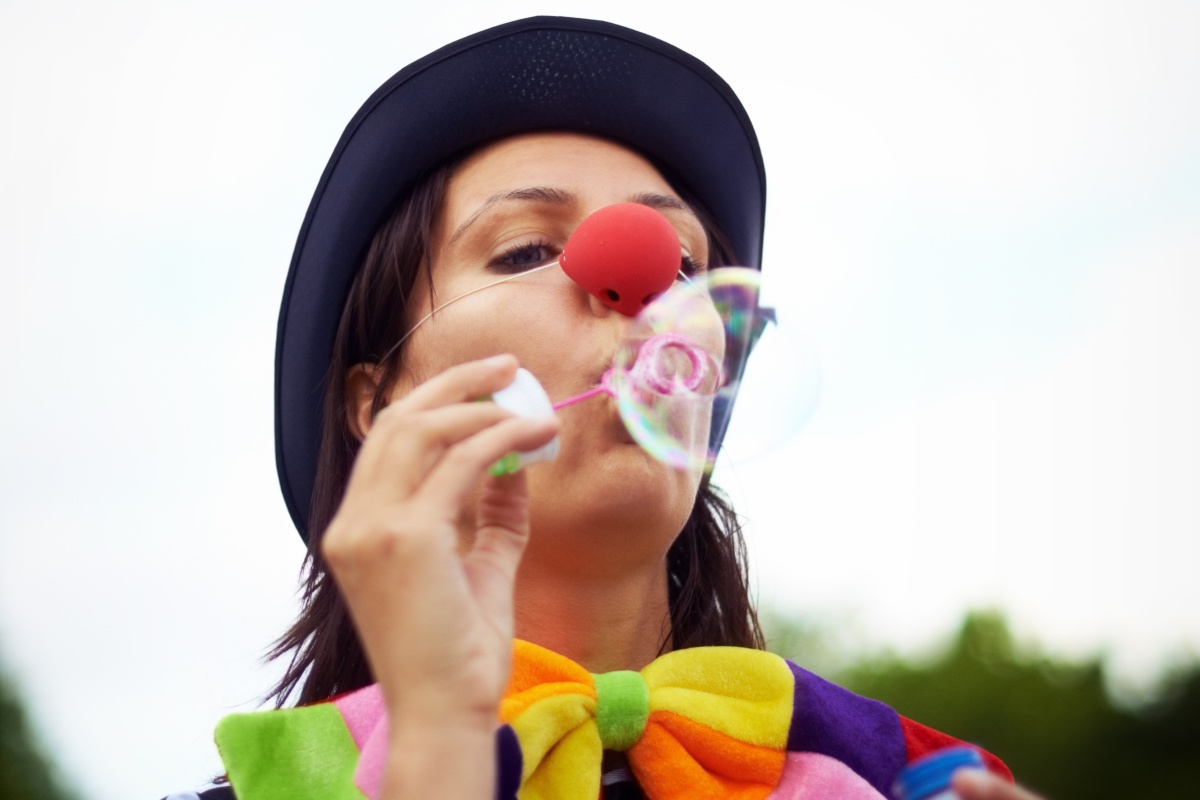 Person mit Clownsnase, Hut und bunter Fliege pustet Seifenblasen in die Luft.