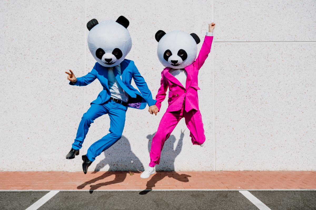 Zwei Personen in blauem und pinkem Anzug mit Panda-Masken springen fröhlich vor einer hellen Wand während sie sich den Händen halten.