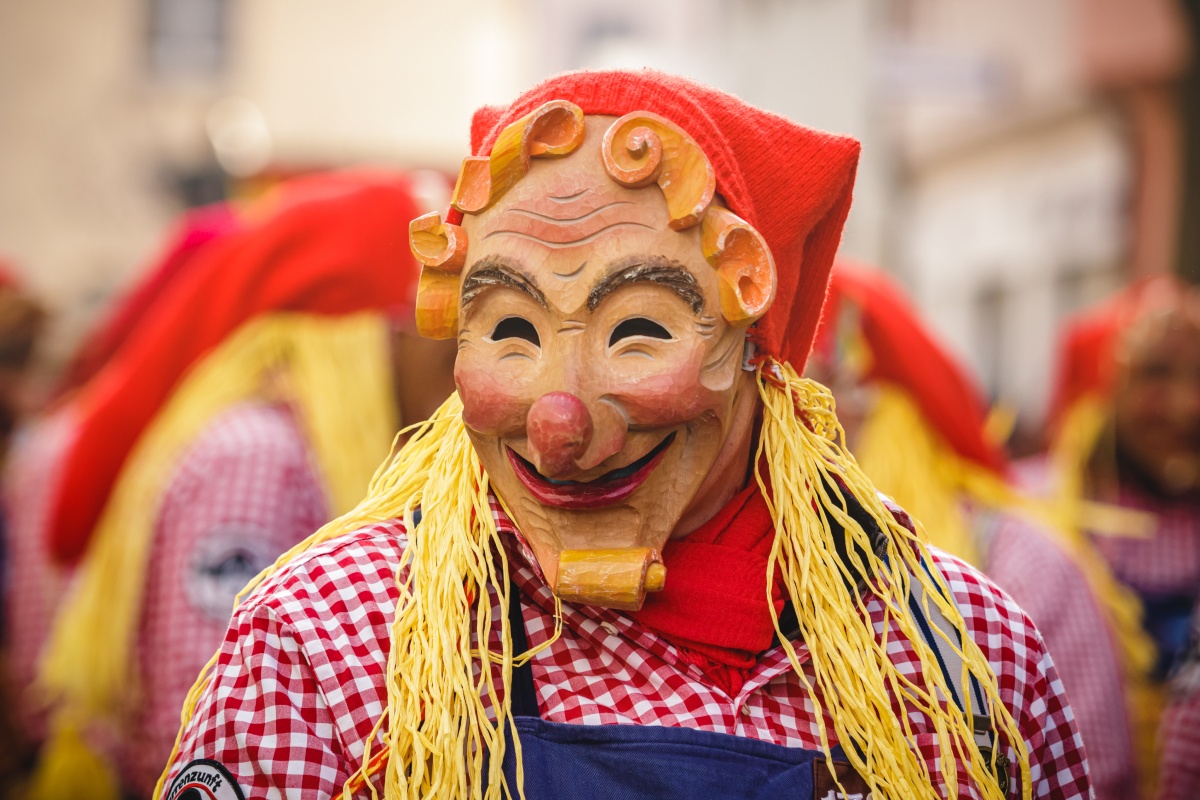 Person in traditioneller Holzmaske, roter Mütze und kariertem Hemd auf einem Karnevalsumzug.