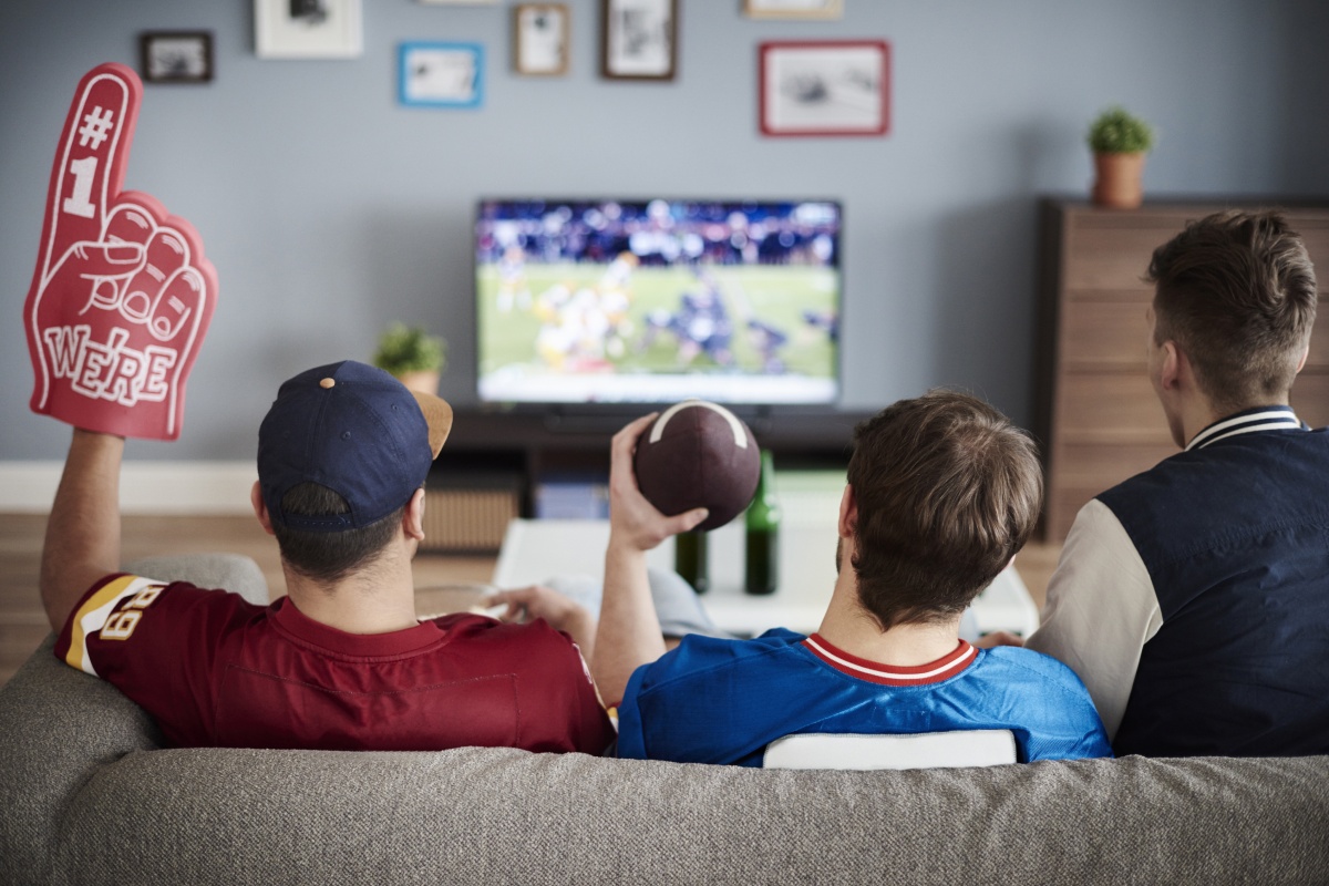 Drei Männer sitzen auf einer Couch im Wohnzimmer und schauen sich ein American-Football-Spiel im TV an.