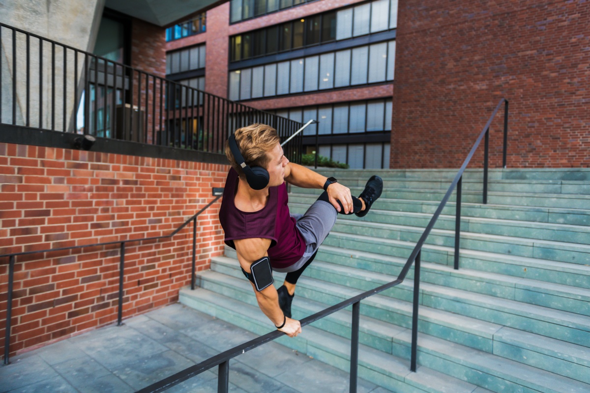 Junger Mann mit großen Kopfhörern spingt über ein Treppengeländer vor einem modernen Bürogebäude.