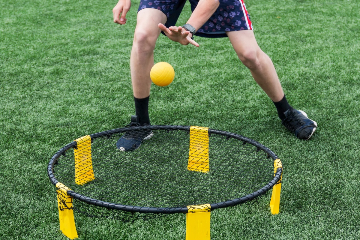 Person in sportlichem Outfit spielt mit einem gelben Ball auf einem Spikeball-Netz.