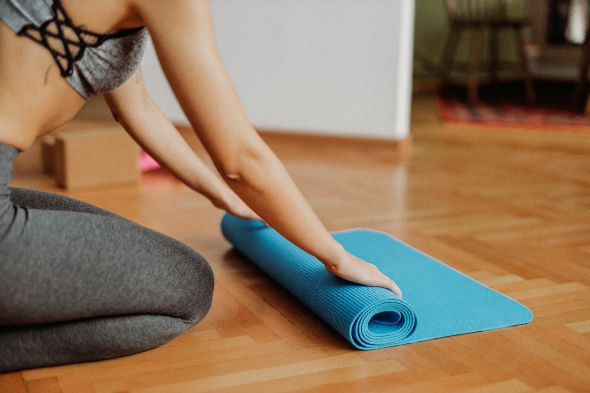 Frau in Sportkleidung rollt eine blaue Yogamatte auf einem Holzboden zusammen.
