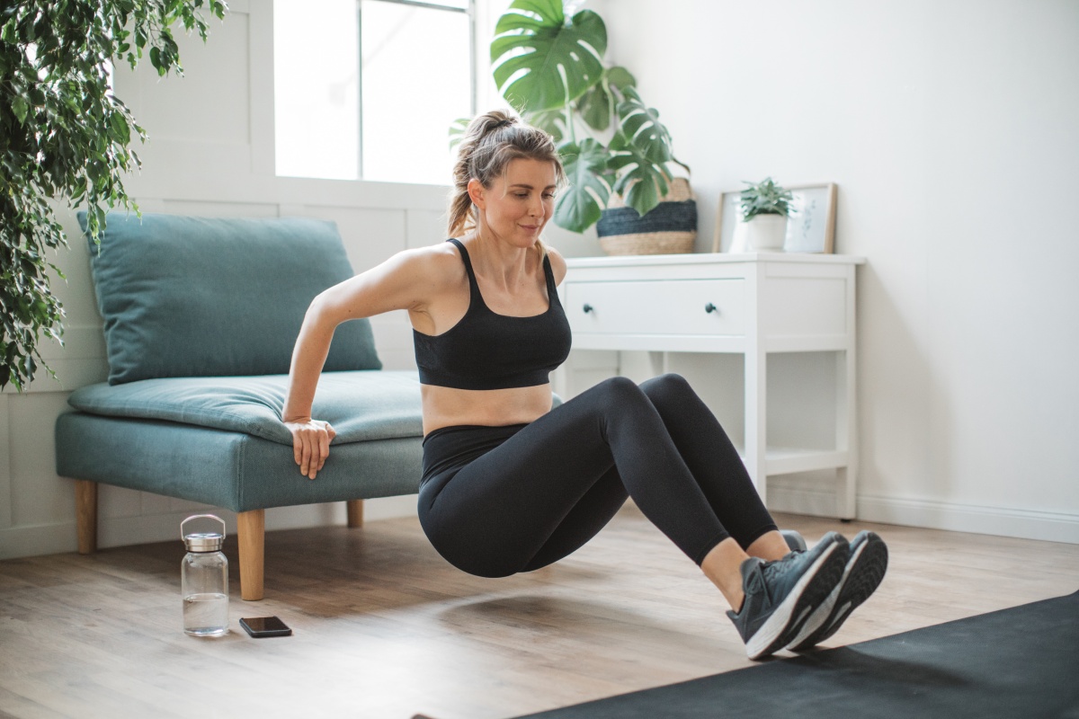 Frau in schwarzer Sportkleidung macht Dips an einem Polsterstuhl zu Hause im Wohnzimmer.