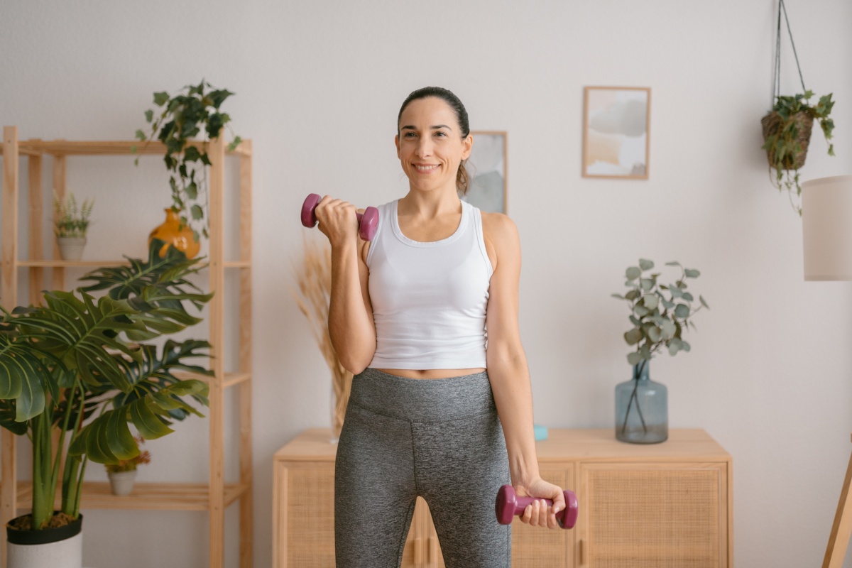Frau in Sportkleidung trainiert mit kleinen Hanteln in einem hell eingerichteten Raum mit Pflanzen und Holzmöbeln.