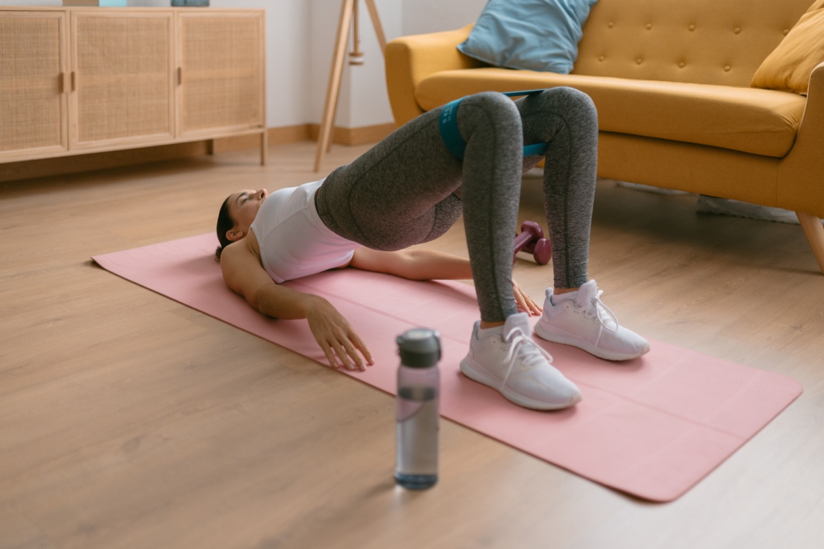 Frau macht Hüftbrücke mit Fitnessband auf rosa Yogamatte in modernem Wohnzimmer.