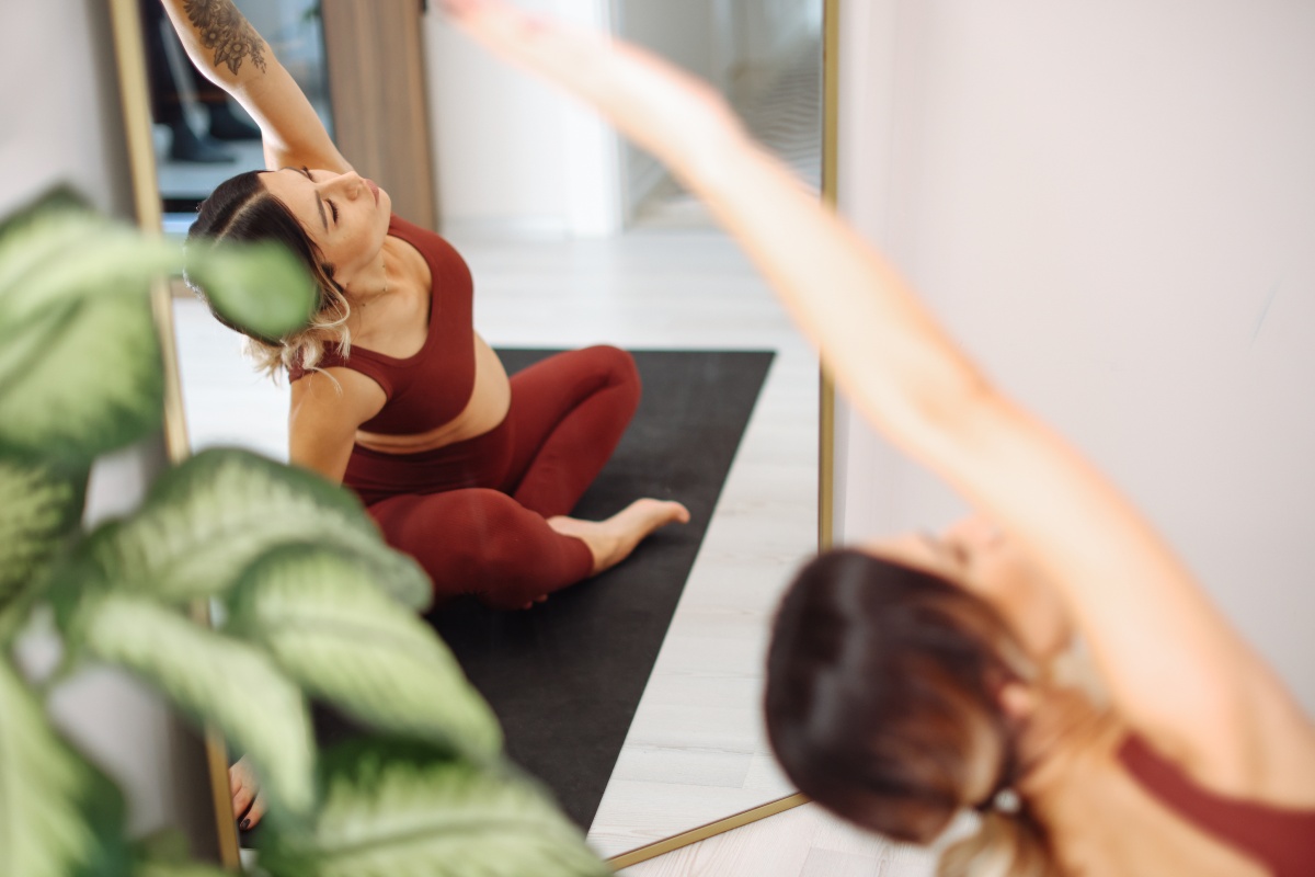 Frau sitzt im Schneidersitz auf einer schwarzen Matte und macht Yoga-Übungen vor einem großen Spiegel.