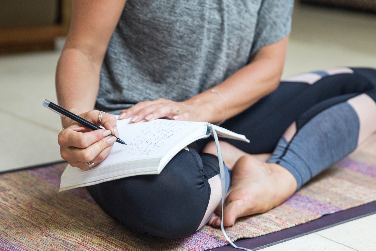 Frau sitzt im Schneidersitz auf Yogamatte und schreibt in ein Notizbuch.