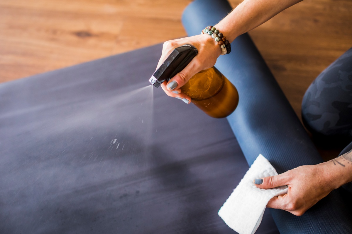 Frau reinigt Yogamatte mit Sprühflasche und Tuch in der Hand.