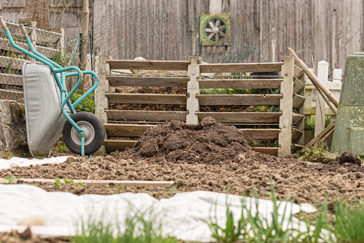 Ein umgegrabenes Beet, dahinter Komposter aus Holz, links davon eine an den Zaun angelehnte Schubkarre.