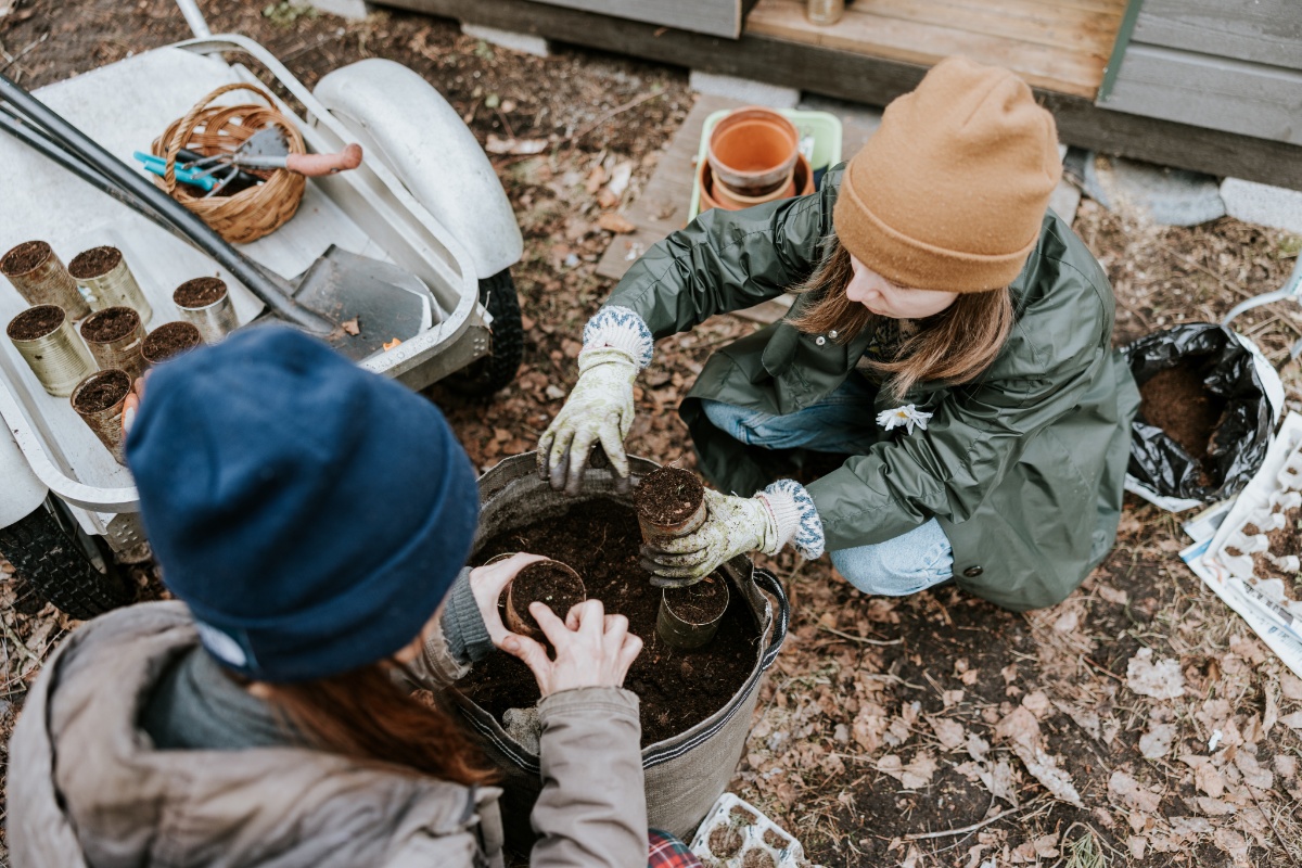 Zwei Personen mit Mütze hocken auf der Erde und befüllen alte Dosen mit Komposterde, um sie herum Gartengeräte, Dosen und Töpfe.