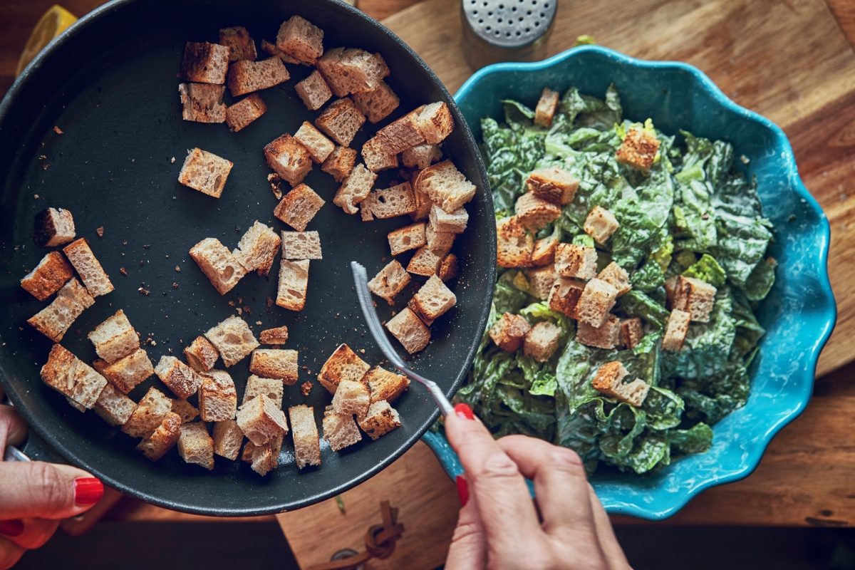 Person gibt geröstete Croutons in einen Feldsalat mit Joghurtdressing.