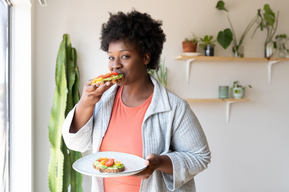 Frau isst ein belegtes Avocado-Toast mit Tomaten und hält einen Teller.