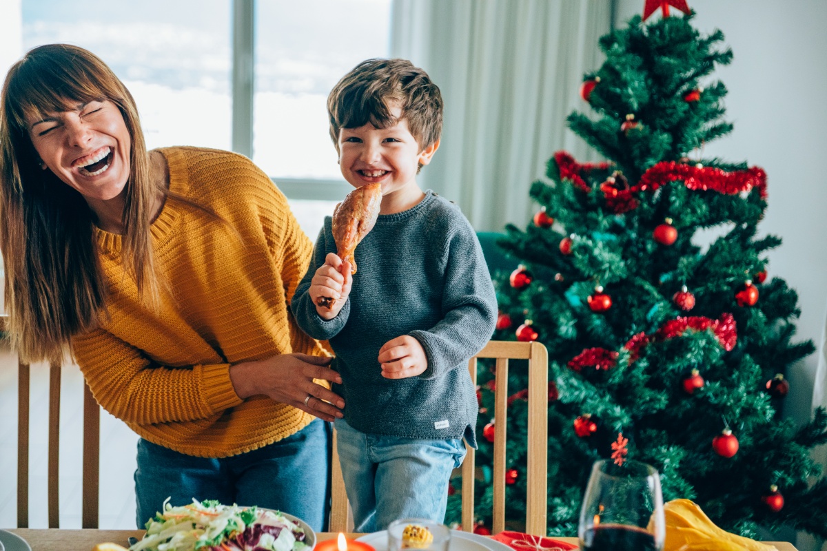 Lachende Mutter mit Kind, das Hähnchenkeule am festlich dekorierten Weihnachtstisch hält.
