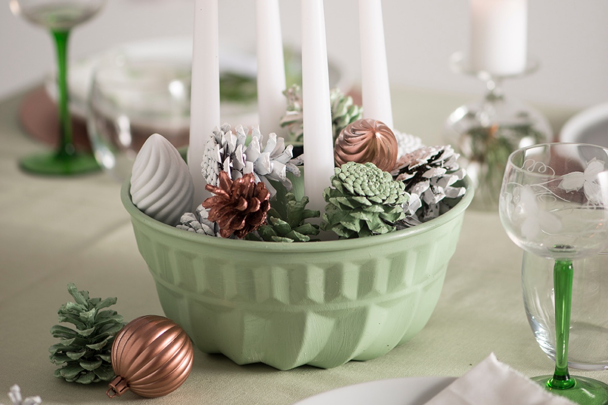 Grünes Dekoschälchen mit weißen Kerzen, bemalten Tannenzapfen und kupferfarbenen Weihnachtskugeln.