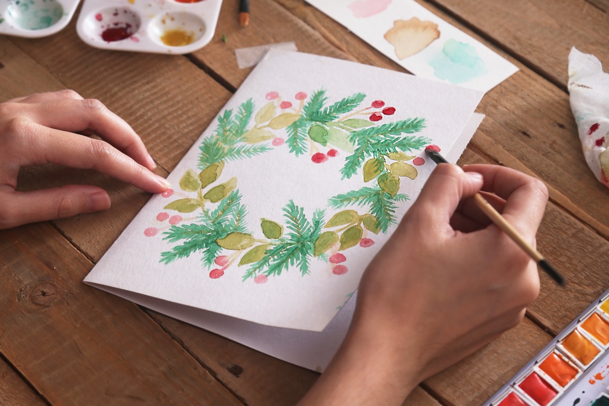 Person malt einen weihnachtlichen Kranz mit Aquarellfarben auf eine Weihnachtskarte.