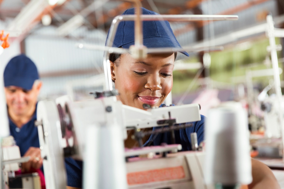 Frau in blauer Kappe arbeitet konzentriert an einer Nähmaschine in einer Fabrik.