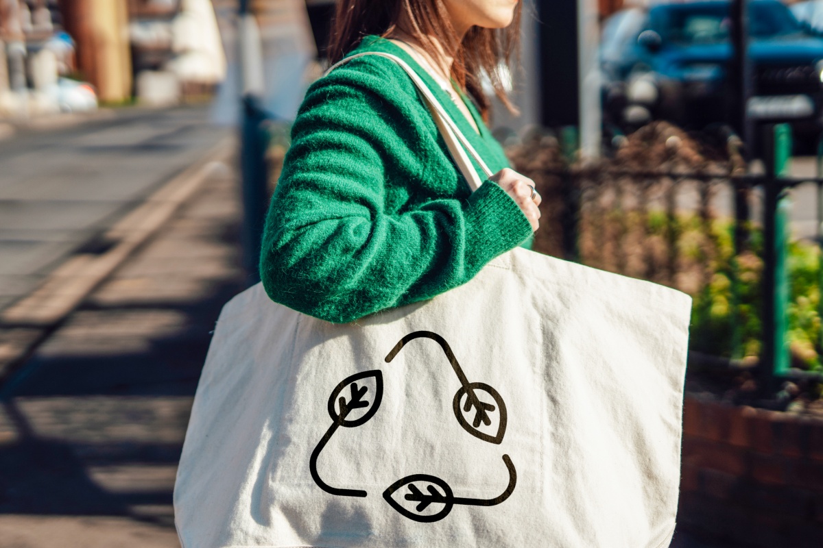 Frau in grünem Pullover trägt eine weiße Stofftasche mit Recycling-Symbol aus Blättern auf der Straße.
