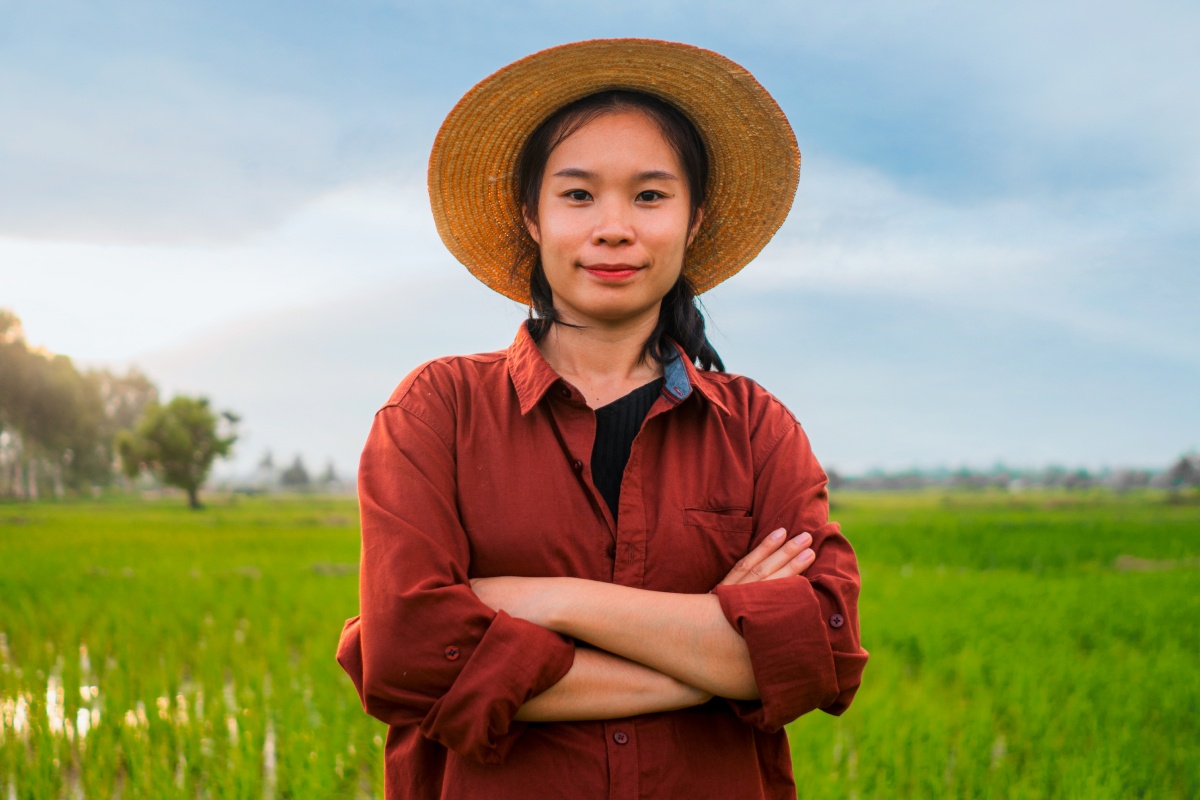 Junge Bäuerin mit Strohhut und rotem Hemd steht auf einem grünen Feld und blickt selbstbewusst in die Kamera.