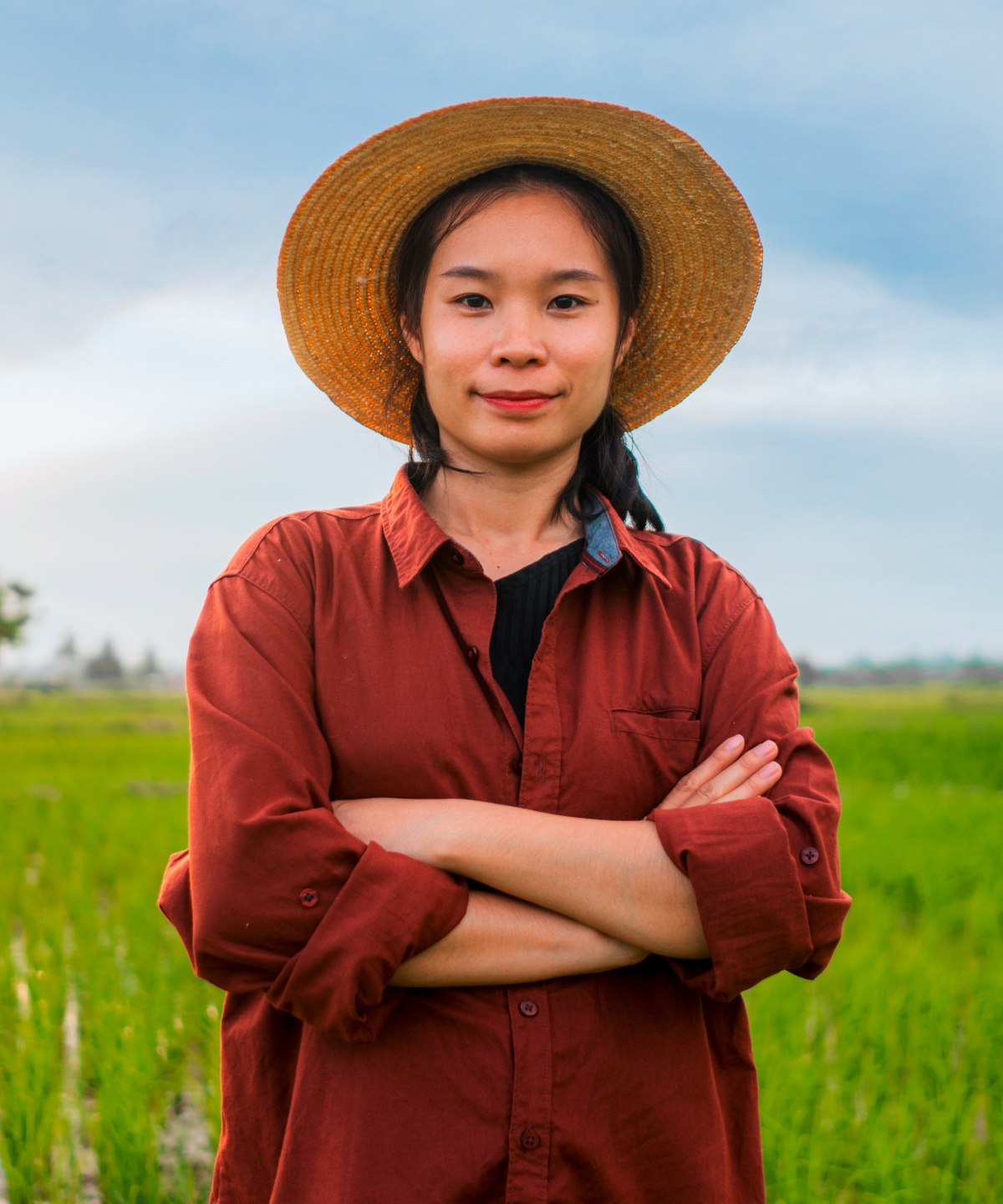 Junge Bäuerin mit Strohhut und rotem Hemd steht auf einem grünen Feld und blickt selbstbewusst in die Kamera.
