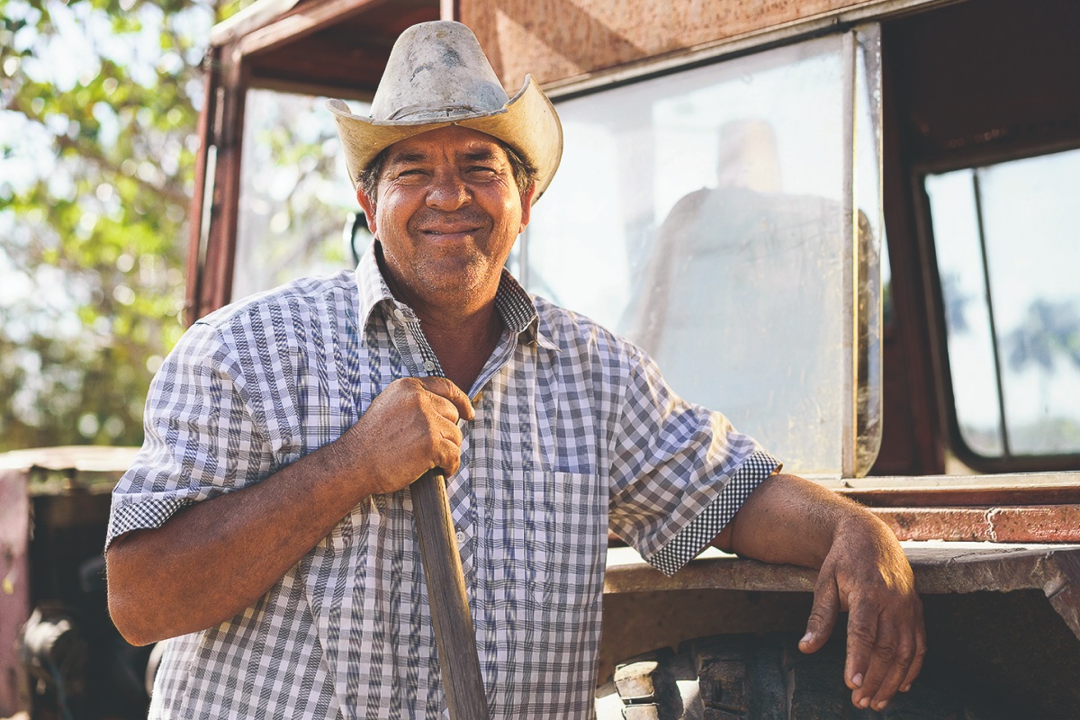 Bauer mit Hut und kariertem Hemd lächelt vor einem Traktor in einer ländlichen Umgebung.