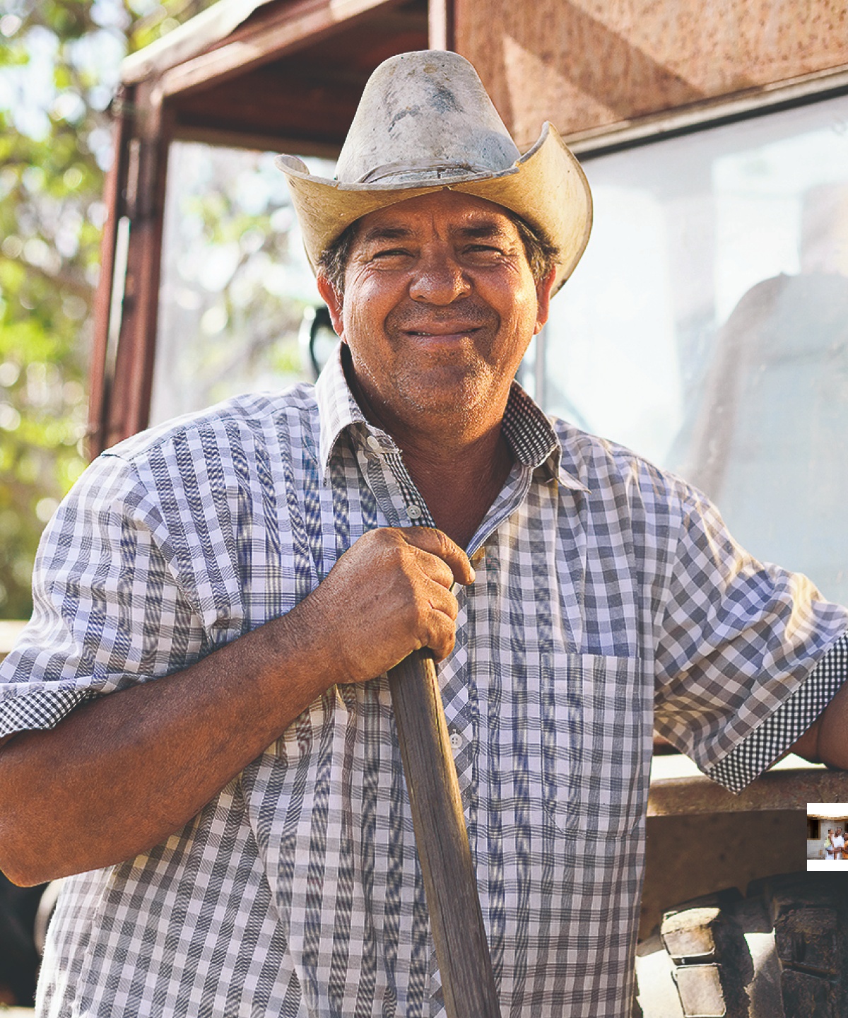 Bauer mit Hut und kariertem Hemd lächelt vor einem Traktor in einer ländlichen Umgebung.