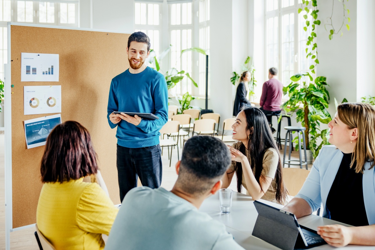 Ein Mann präsentiert einem kleinen Team in einem modernen Büro, umgeben von Pflanzen und großen Fenstern, Diagramme auf einem Whiteboard.