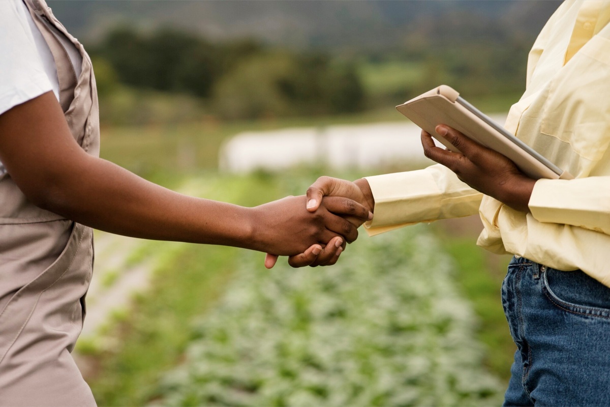 Zwei Personen schütteln sich die Hand auf einem Feld. Eine Person hält ein Tablet.