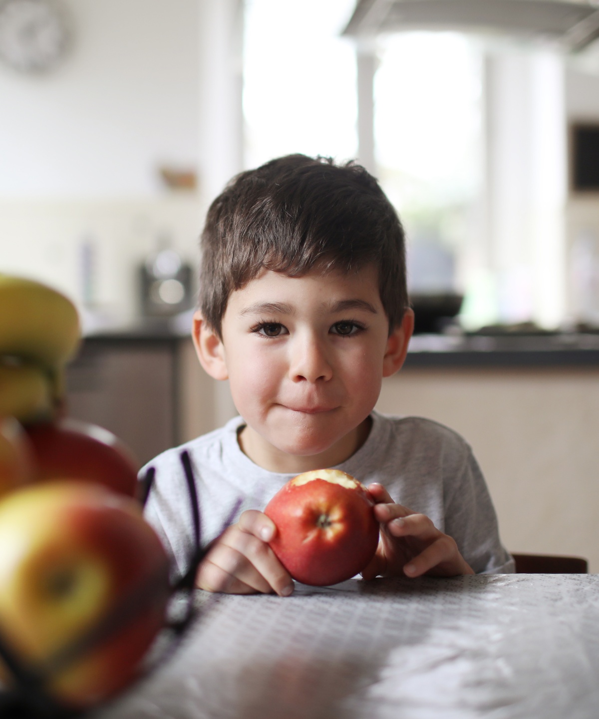 Junge isst einen Apfel am Küchentisch, vor einem Obstkorb.