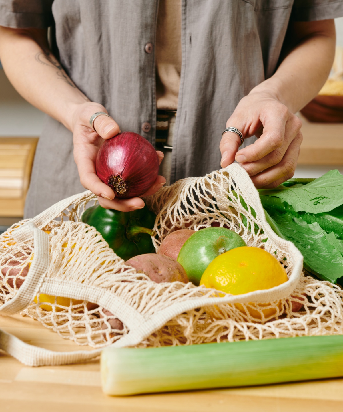 Person packt frisches Gemüse und Obst, darunter rote Zwiebel, Paprika und Avocados, aus einem Netzbeutel aus.