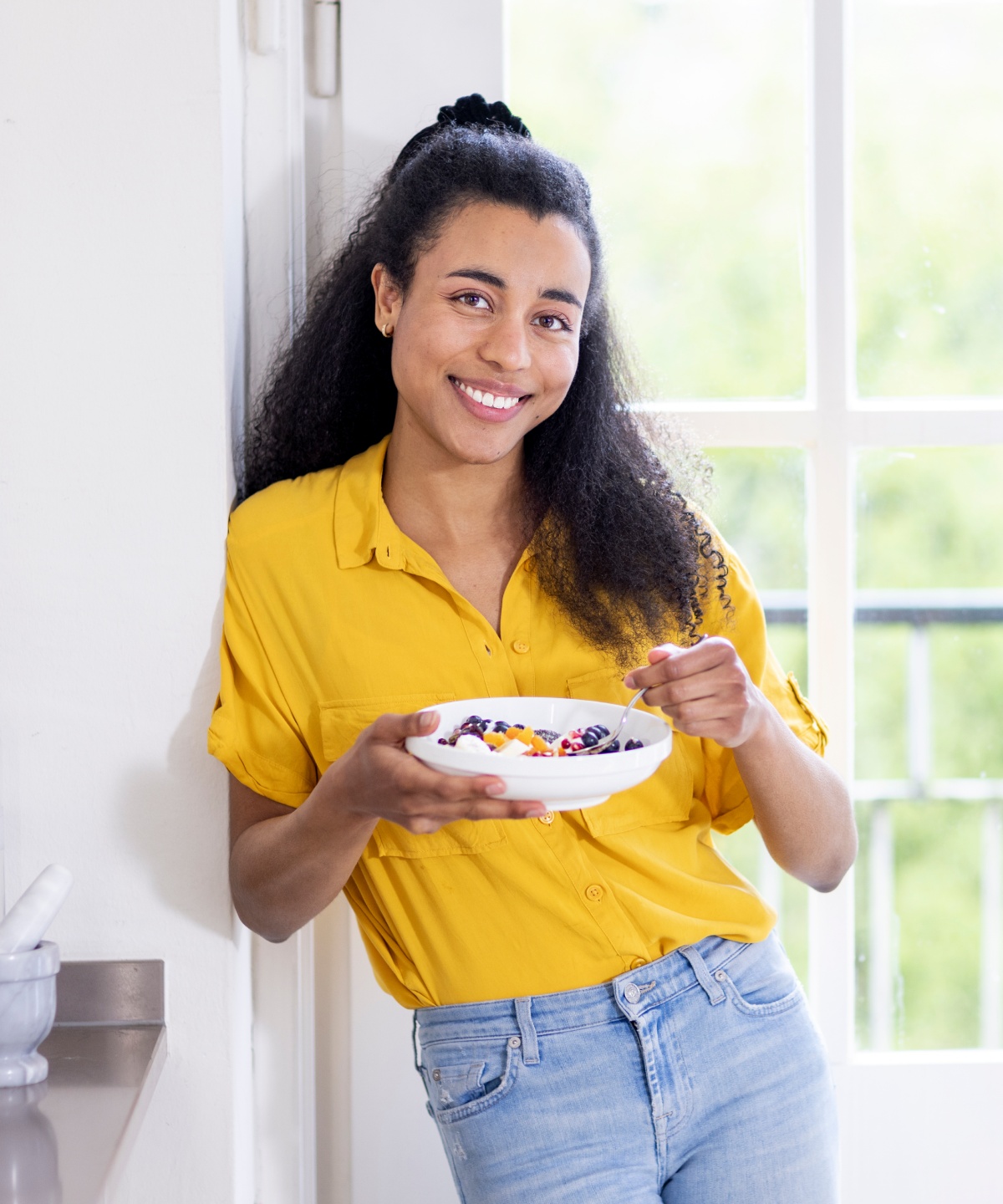 Lächelnde Frau in gelber Bluse steht mit einer Frühstücksschale vor einem hellen Fenster.