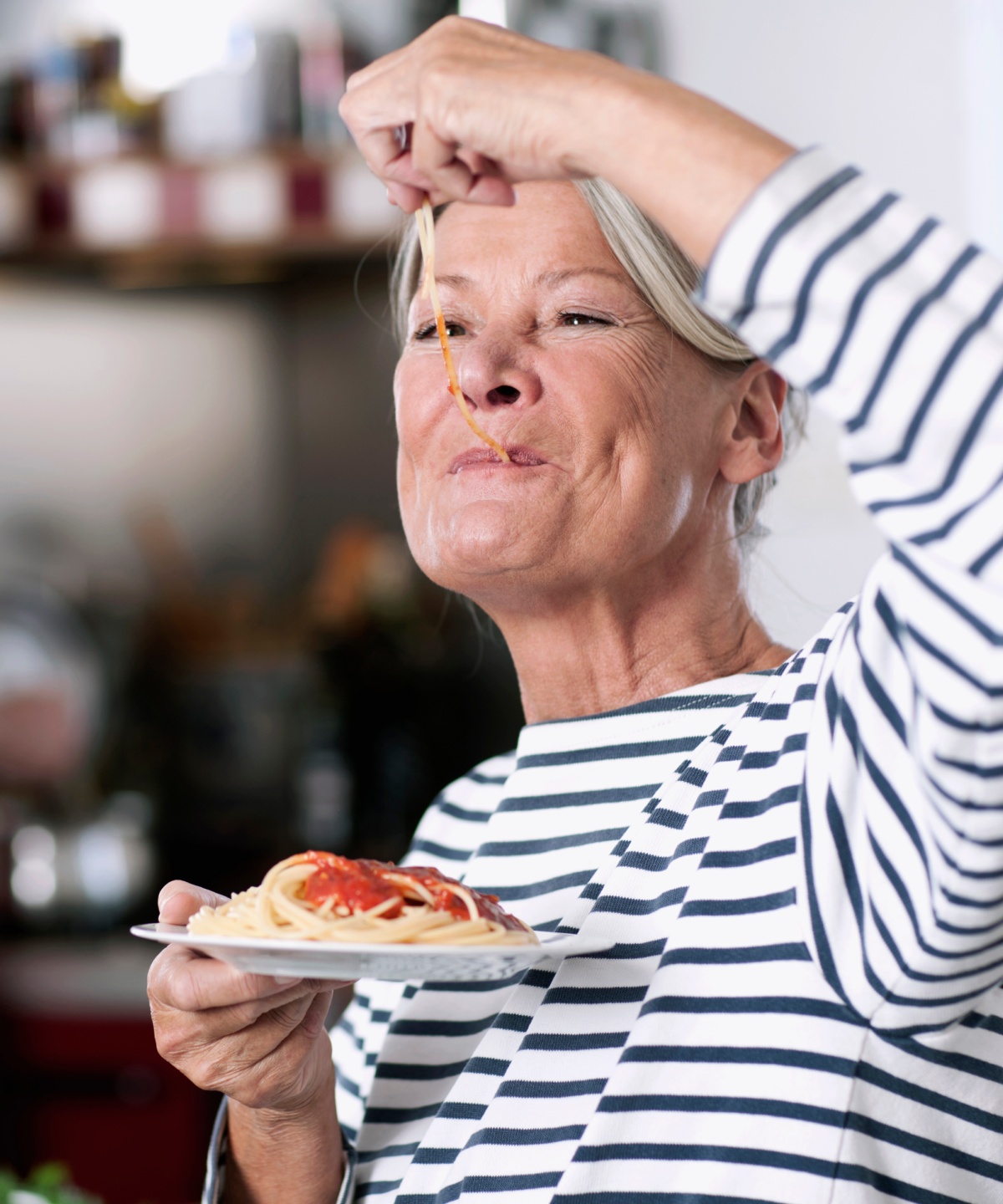 Ältere Frau genießt Spaghetti in der Küche, im Hintergrund kocht ein älterer Mann.