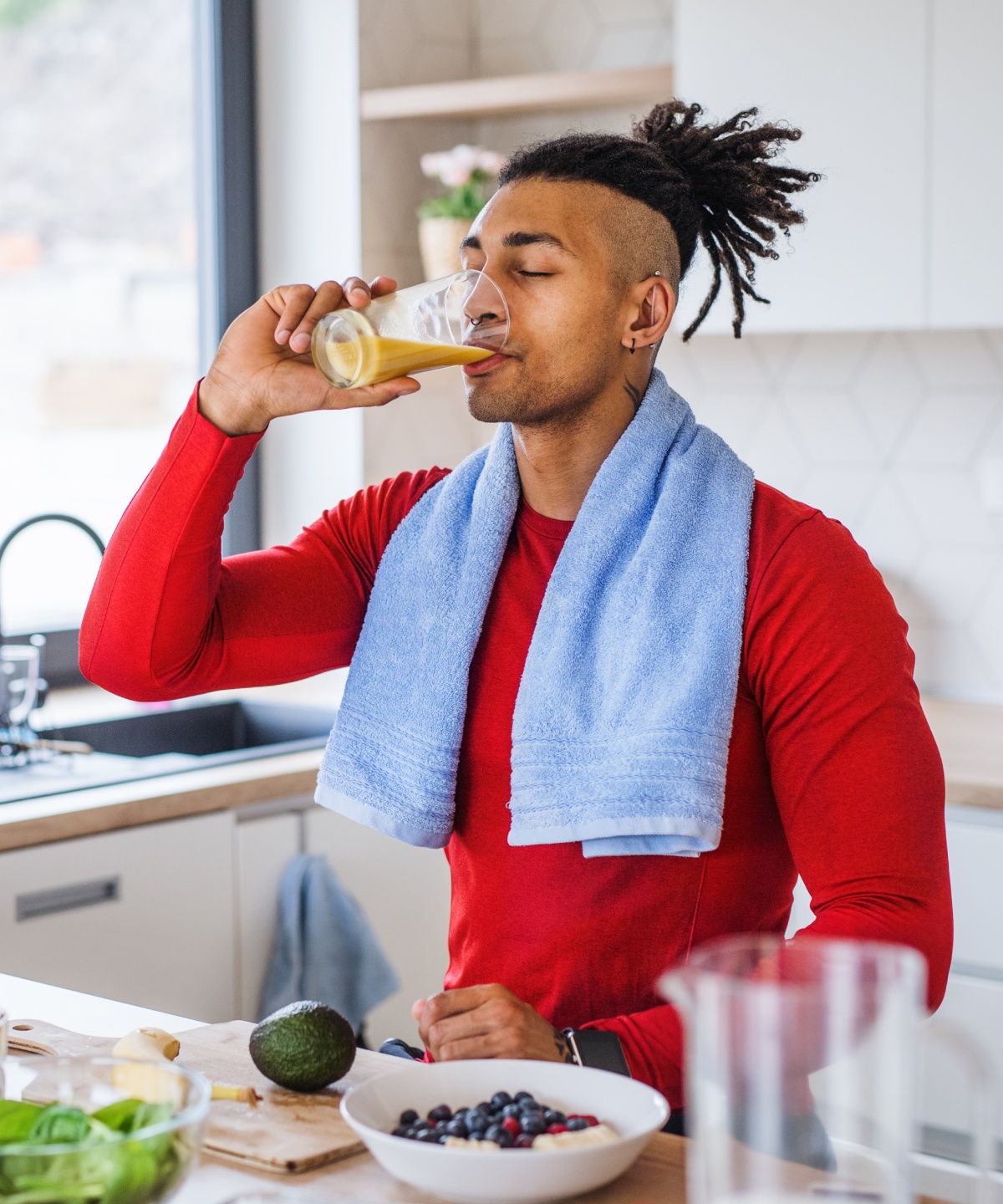 Sportlicher Mann in rotem Shirt trinkt Saft in einer modernen Küche, ein Handtuch um den Hals, Obst und Gemüse auf der Theke.