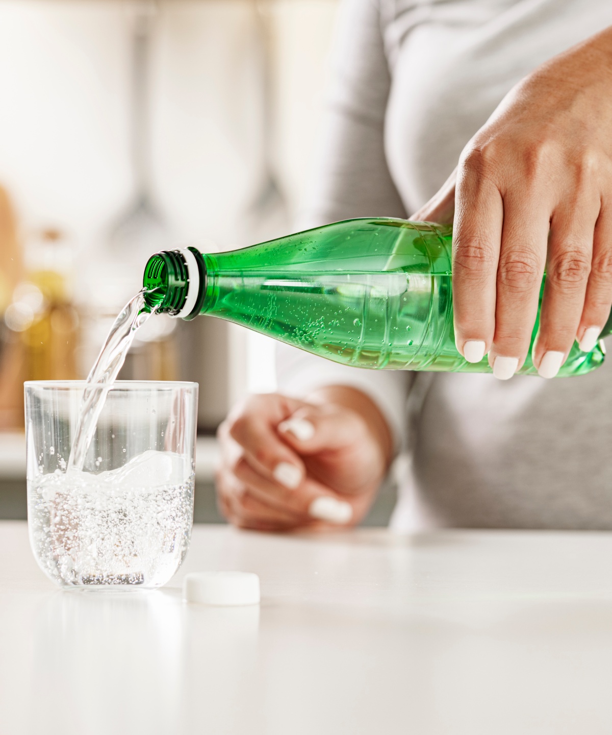 Person gießt Mineralwasser aus einer grünen PET-Flasche in ein Glas, das auf einer Küchenarbeitsplatte steht.