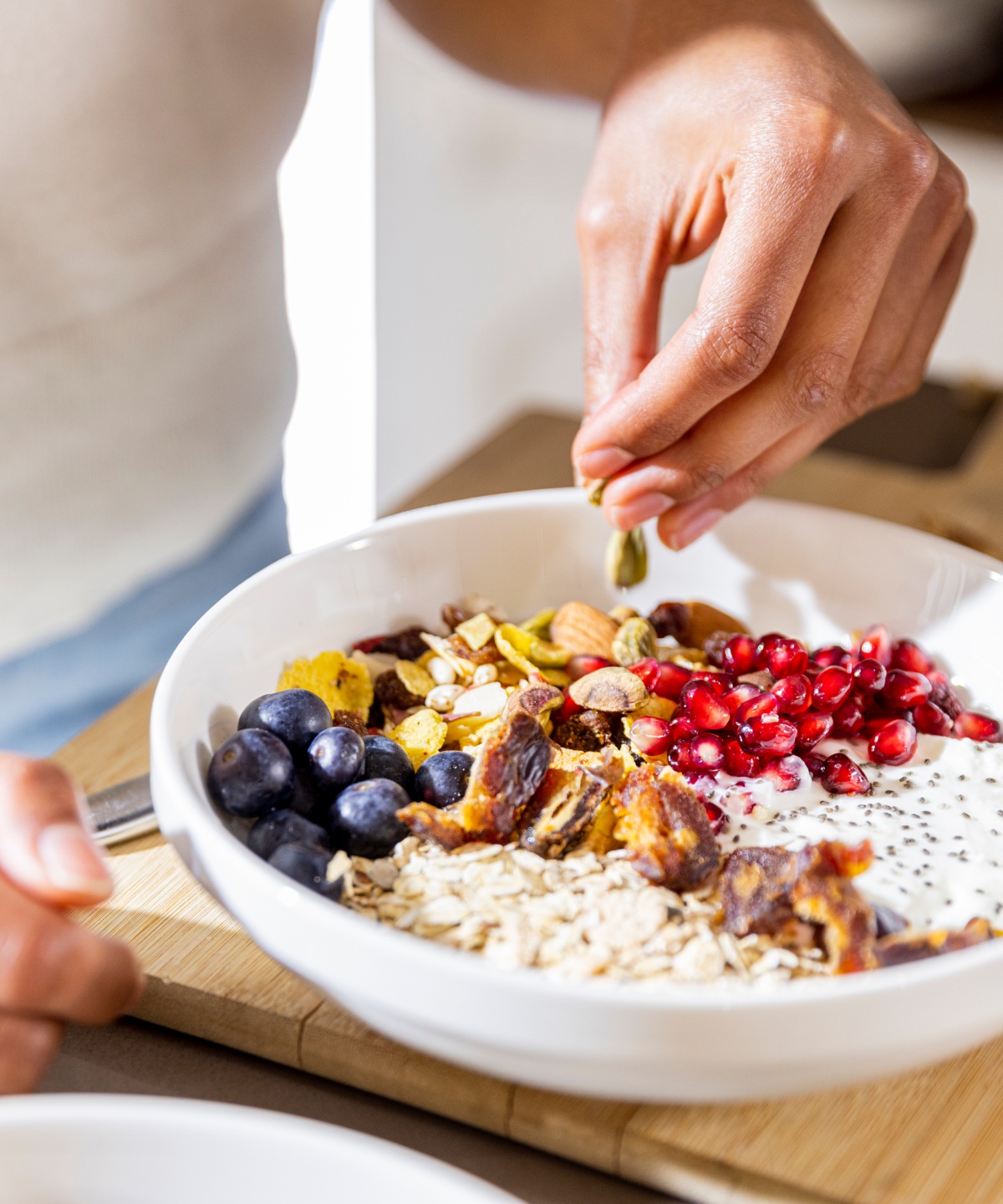 Person garniert eine Frühstücksschale mit Haferflocken, Blaubeeren, Granatapfelkernen, Nüssen und Joghurt auf einem Holztisch.