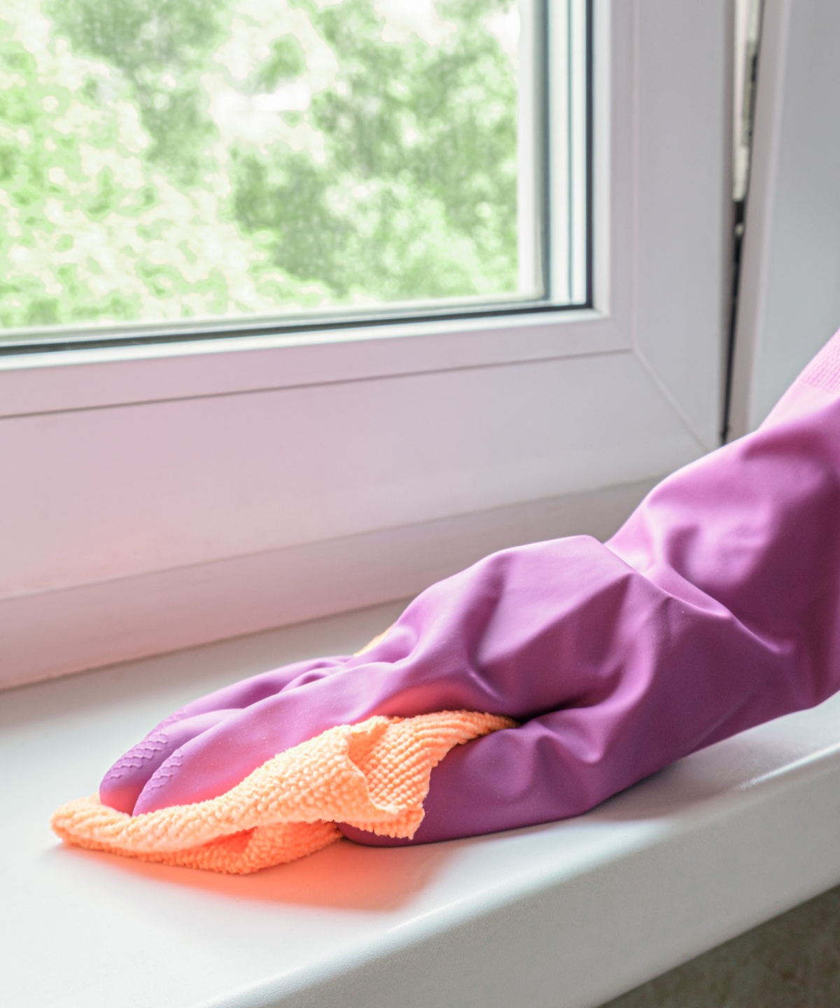 Eine Person, die Putzhandschuhe trägt, wischt Schmutz mit einem Lappen von der Fensterbank.