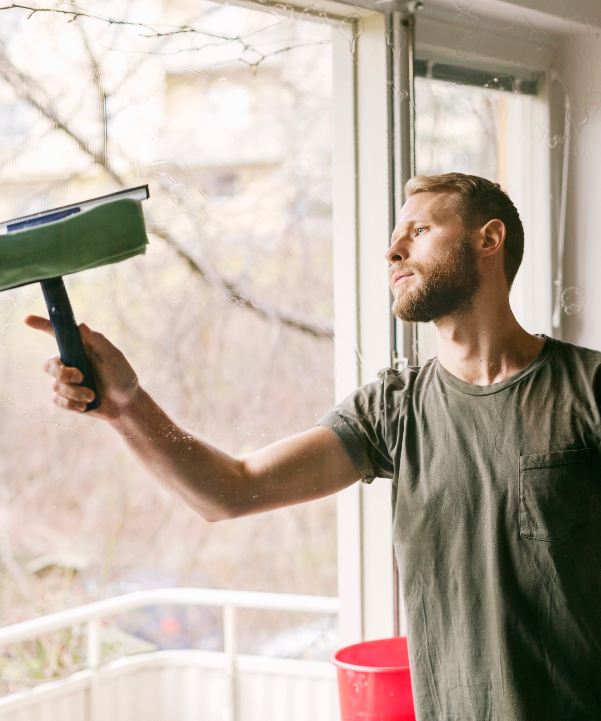 Ein Mann putzt sein Balkonfenster mit einem Abzieher. Neben ihm steht ein Eimer mit Putzwasser.