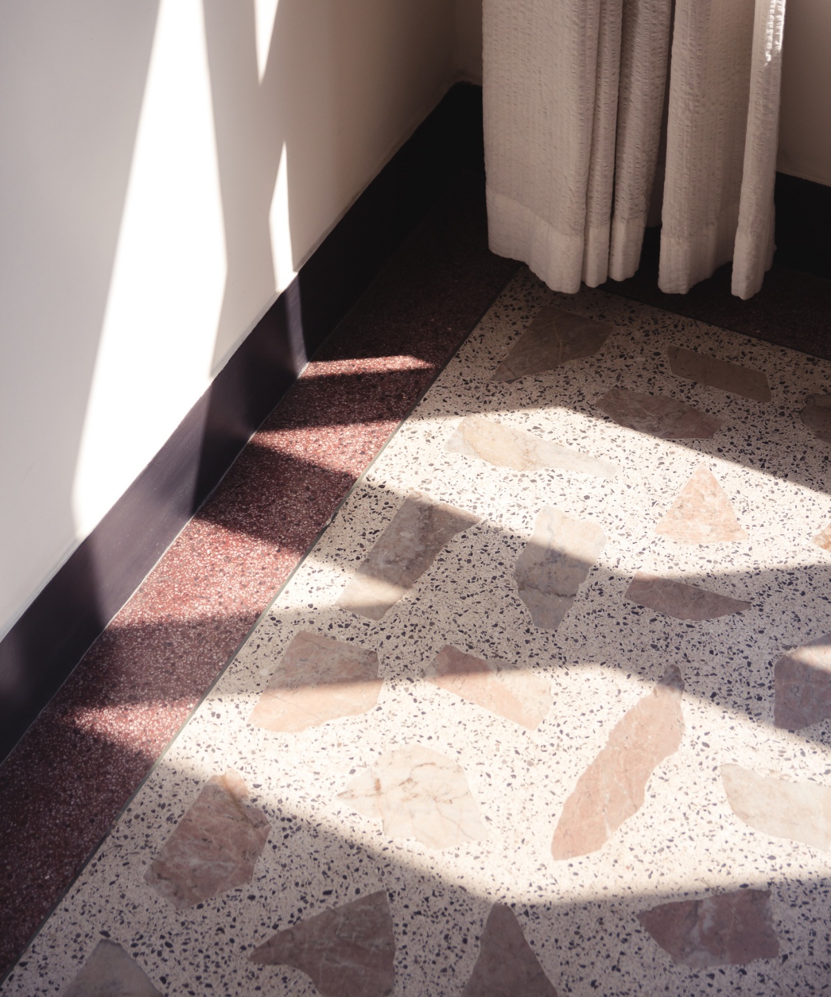 Fußboden im Terrazo-Stil, Schatten fallen auf den Boden und die Wand, unterer Teil eines Vorhangs ist zu erkennen.
