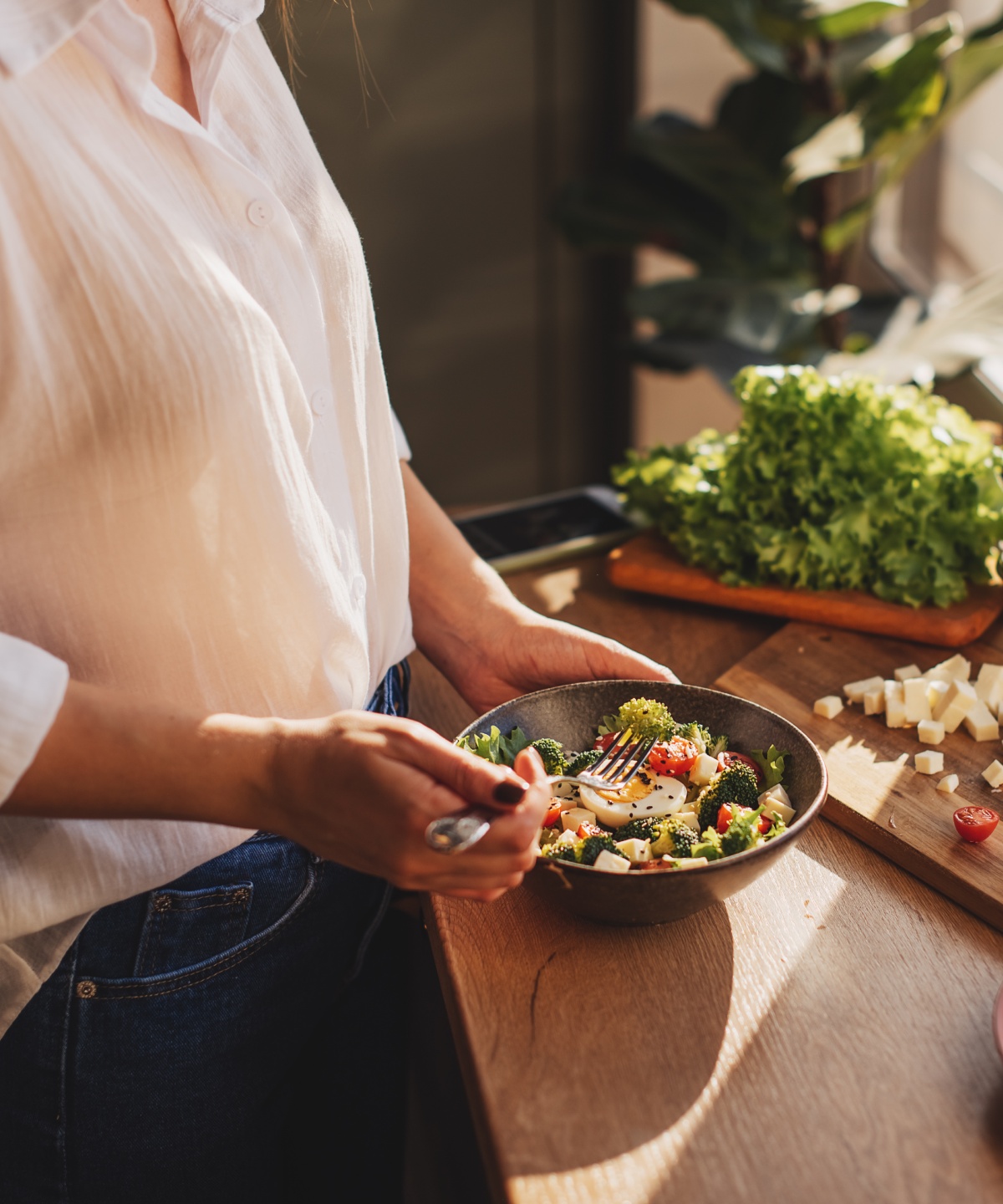 Frau bereitet in einer sonnigen Küche einen frischen Salat mit Brokkoli, Tomaten, Käse zu.