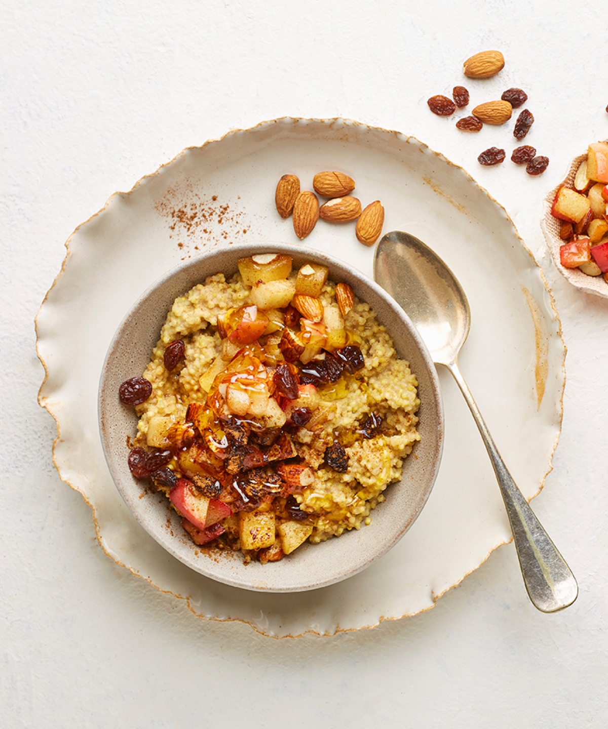 Schüssel mit Hirsebrei, garniert mit Äpfeln, Mandeln und Rosinen, daneben frischer Apfel und Löffel.