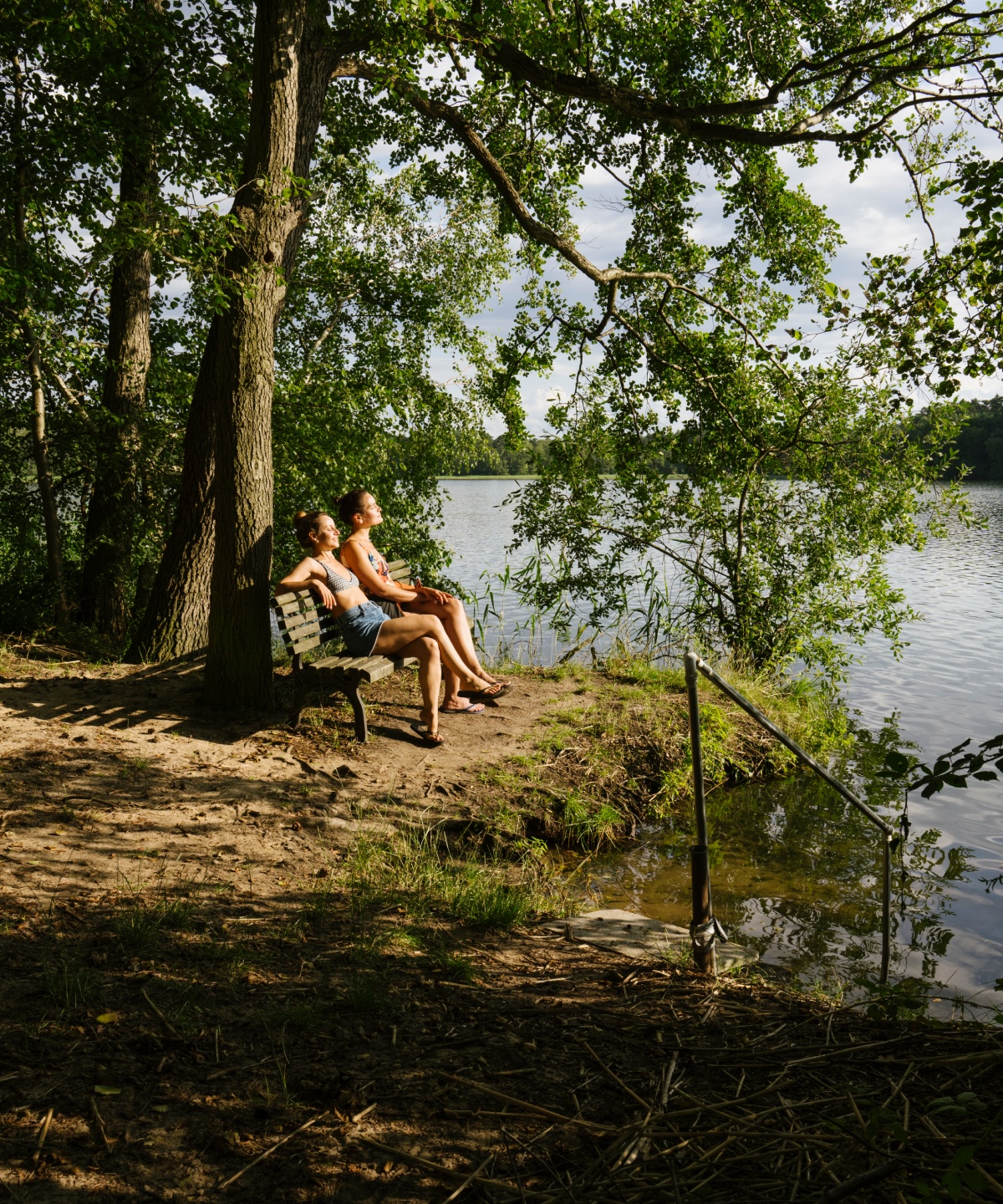Zwei Frauen sitzen auf eine Holzbank an einem Waldsee, den Blick in die Sonne gerichtet.