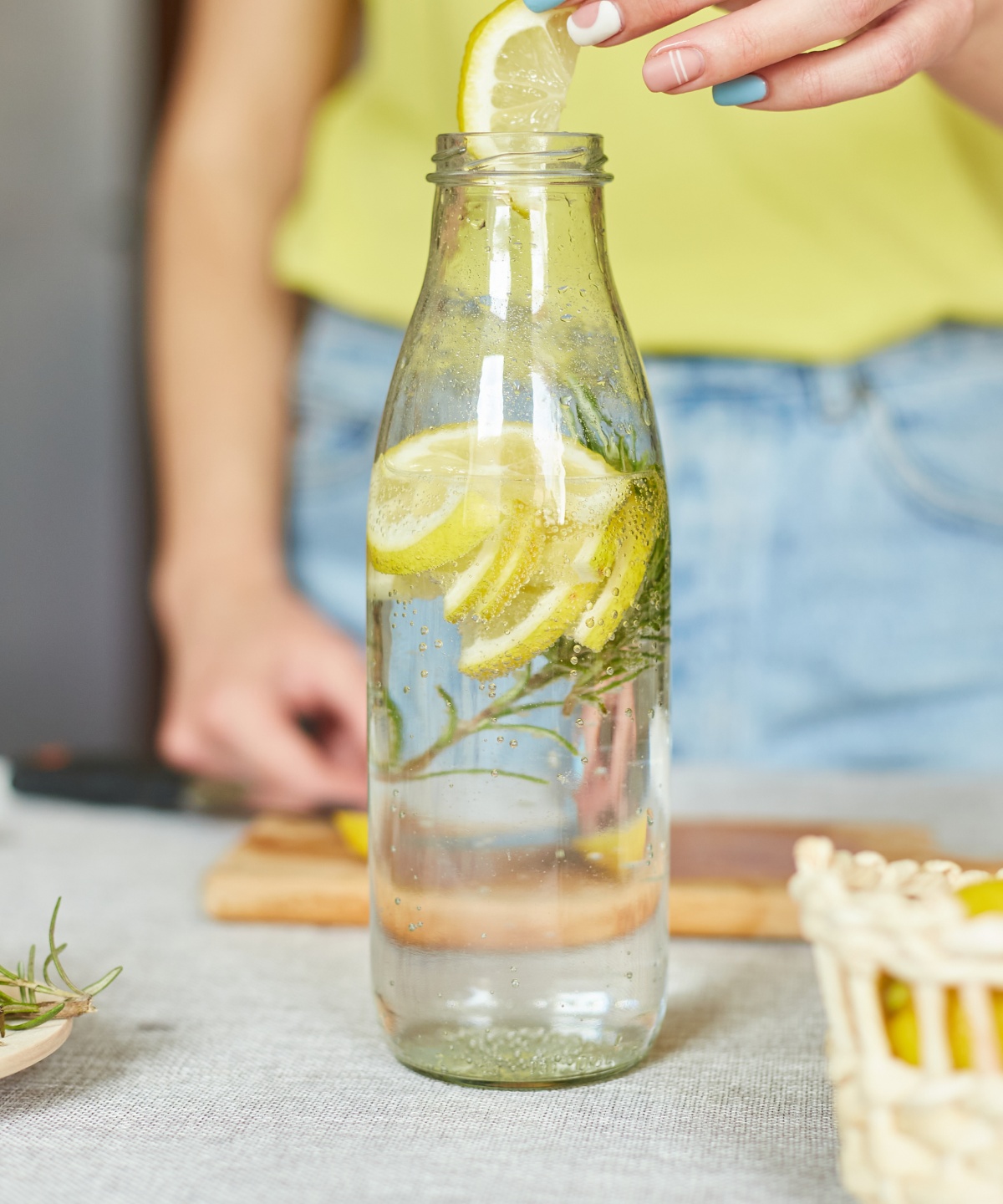 Person fügt Zitronenscheibe in eine Flasche Wasser mit Rosmarin, daneben Zitronen und ein Korb.