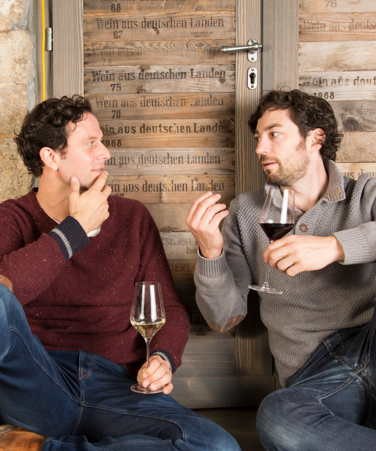 Zwei Winzer unterhalten sich über die Qualität von Wein. Dabei sitzen sie vor einer Holztür mit einem Glas Wein in der Hand.