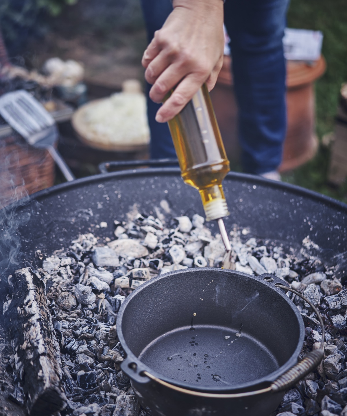 Person gießt Öl aus einer Flasche in einen Dutch Oven-Topf, der in einer mit Kohle erhitzten Feuerschale steht. Im Hintergrund unscharf Garten und Grillzubehör.